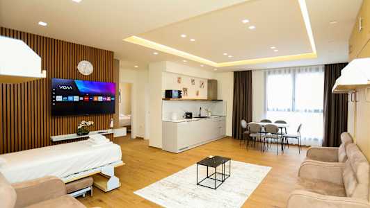 Et moderne hotelværelse med åbent gulvplan, der inkluderer en sovesofa, TV, køkkenområde, spisebord og store vinduer med gardiner. (AI-genereret tekst)