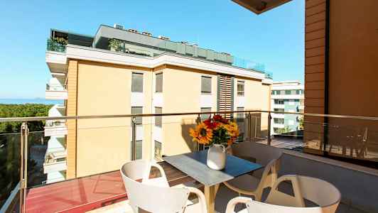 En balkon med udsigt til bygninger og havet. På balkonen er der et bord med en vase med blomster. (AI-genereret tekst)