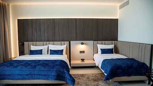 Billede af et hotelværelse med to senge med blå sengetæpper og hvide puder. Farverige tæpper ses på gulvet. (AI-genereret tekst)