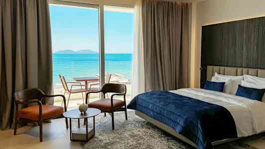 Et hotelværelse med havudsigt med en dobbeltseng, to stole, et lille bord og en glasdør til en terrasse. (AI-genereret tekst)