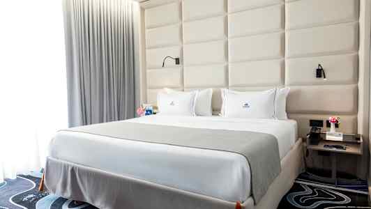 Et soveværelse med en stor seng, hvide lagen og et gråt sengetæppe. Bag sengen er der en polstret senggavl, og på siderne står natborde med lamper. (AI-genereret tekst)