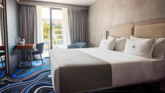 Et hotelværelse med en opredt dobbeltseng, et skrivebord med stol og vindue mod altan. (AI-genereret tekst)