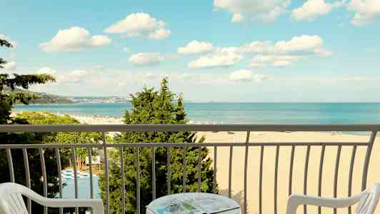 Udsigt fra en altan mod en strand og havet, med stole og et bord i forgrunden. (AI-genereret tekst)