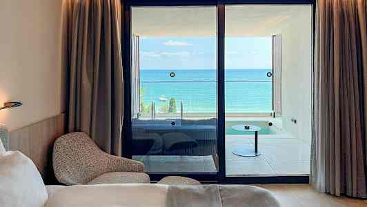 Et hotelværelse med en seng og lænestol, samt udsigt til en balkon med havet i baggrunden. (AI-genereret tekst)