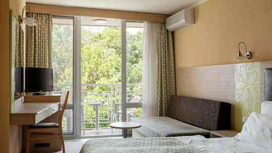 Et hotelværelse med seng, skrivebord, stol og sofa. Vindue og altan med udsigt til grønne områder. (AI-genereret tekst)
