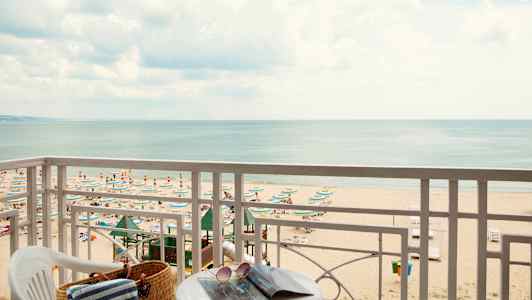 Udsigt fra balkon mod en strand med parasoller og solsenge samt havet i baggrunden. (AI-genereret tekst)
