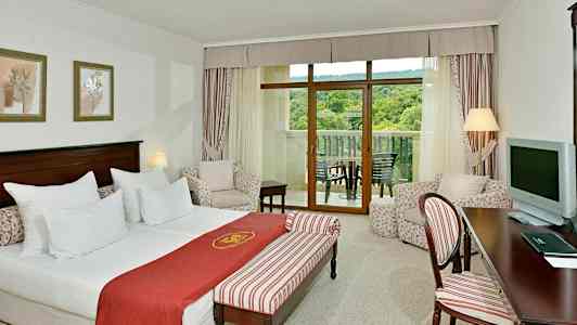 Hotelværelse med seng, siddegruppe og altan med udsigt til grønt. (AI-genereret tekst)