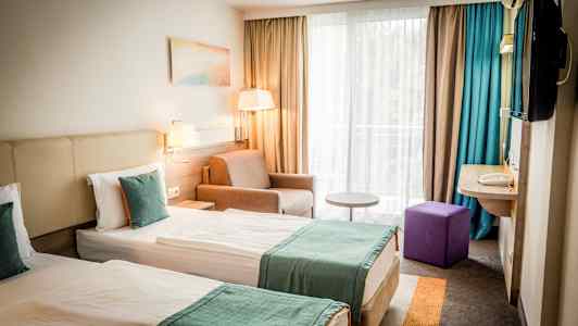 To enkeltsenge med turkise tæpper i et hotelværelse med vindue og siddeområde. (AI-genereret tekst)