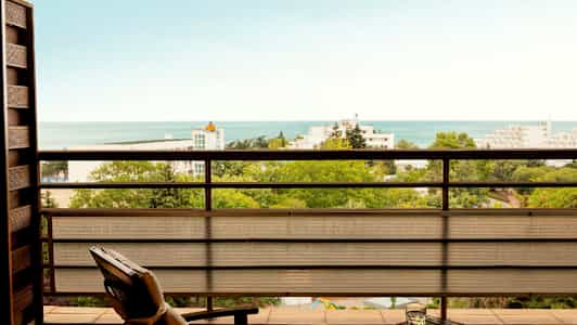 Udsigt fra en altan over grønne områder, bygninger og havet. (AI-genereret tekst)