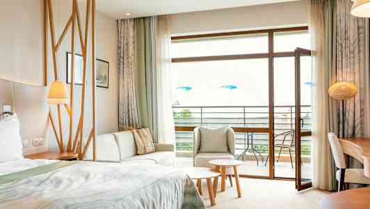 Et hotelværelse med seng, siddeområde, bord og åben altandør. (AI-genereret tekst)