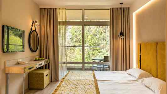 Et hotelværelse med en dobbeltseng, gul sengegavl, lille skrivebord og en stor vinduesfront mod naturen. (AI-genereret tekst)