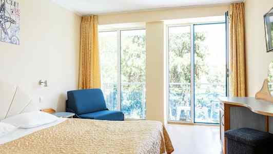 Værelse med seng, blå lænestol og skrivebord, med store vinduer ud mod altan. (AI-genereret tekst)