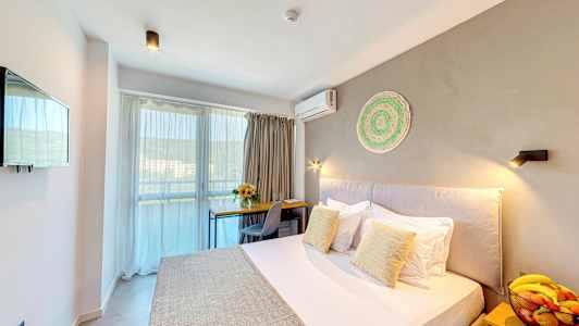 Et hotelværelse med en seng, et skrivebord, en stol, en skål med frugt på et natbord og en altan i baggrunden. (AI-genereret tekst)