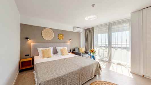 Et lyst hotelværelse med en stor seng, grå sofa og altan med udsigt. (AI-genereret tekst)