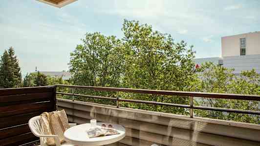 En altan med et bord, to plaststole, en taske, solbriller og en avis samt udsigt til træer og bygninger. (AI-genereret tekst)