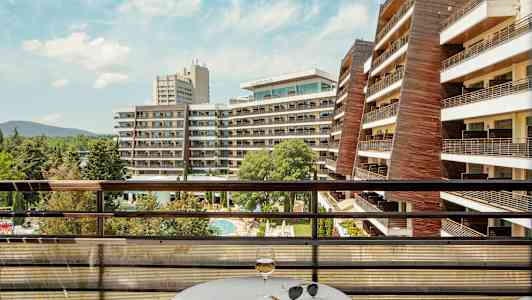 Udsigt fra en altan med bord og glas over hotelbygninger og poolområde. (AI-genereret tekst)