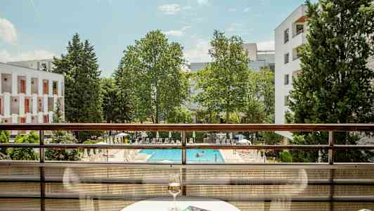 Udsigt fra en altan med udsigt over en pool og grønne områder, med et bord med et glas vin og en bog. (AI-genereret tekst)