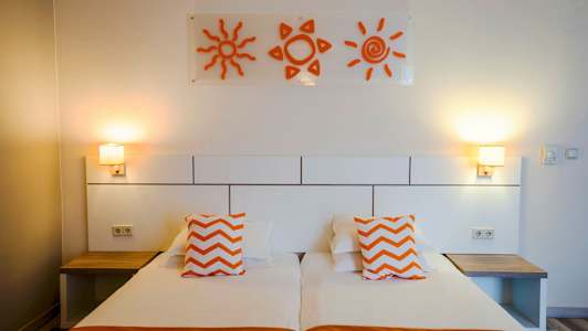 Et hotelværelse med to enkeltsenge, orange puder og vægdekorationer i form af sole. (AI-genereret tekst)