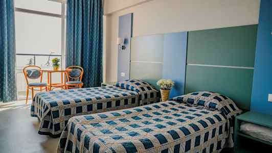 To enkeltsenge i et værelse med blå og grå indretning. Et lille bord med to stole står ved et stort vindue. (AI-genereret tekst)