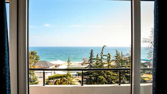 Udsigten gennem et vindue viser havet, nåletræer, huse og en altan. (AI-genereret tekst)