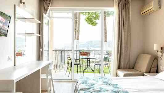 Et hotelværelse med en seng, et bord og en stol samt en balkon med havemøbler og udsigt til naturen. (AI-genereret tekst)