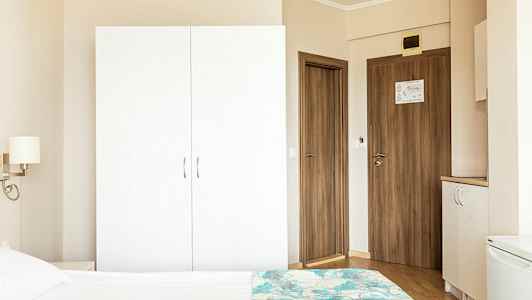 Et hotelværelsesmiljø med et hvidt skab, trædøre og en del af en seng i forgrunden. (AI-genereret tekst)