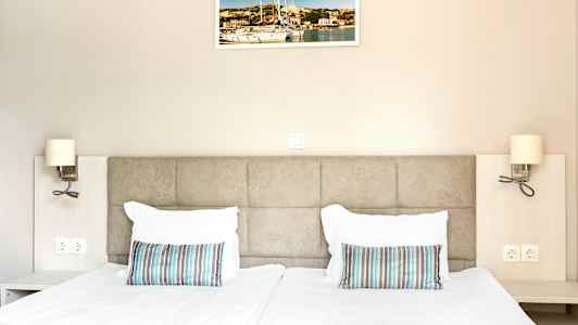 To senge med hvidt sengetøj og stribede puder, væggen bagved har et billede og sengelamper. (AI-genereret tekst)