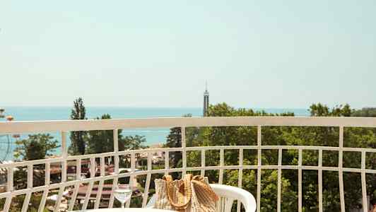 Udsigt fra en balkon med et bord, stole, en taske og et glas vand samt grønne træer og havet i baggrunden. (AI-genereret tekst)
