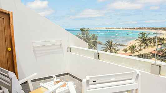 Billedet viser en terrasse med hvide stole og bord samt udsigt over en strand med palmer, hav og små både. (AI-genereret tekst)