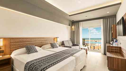 Et hotelværelse med to senge og en siddegruppe ved siden af en glasdør, der fører til en balkon med udsigt. (AI-genereret tekst)