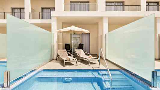 Et poolområde med to liggestole og en parasol foran en hvid bygning med altaner. (AI-genereret tekst)