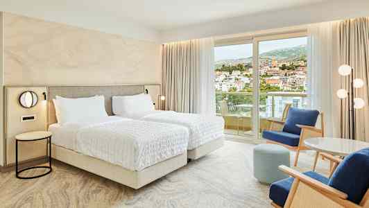 Hotelværelse med to enkeltsenge, siddeområde og balkon med udsigt over en by. (AI-genereret tekst)