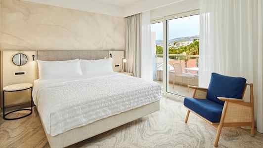 Soveværelse med en stor seng og en blå stol tæt på glasdøre til altanen. (AI-genereret tekst)