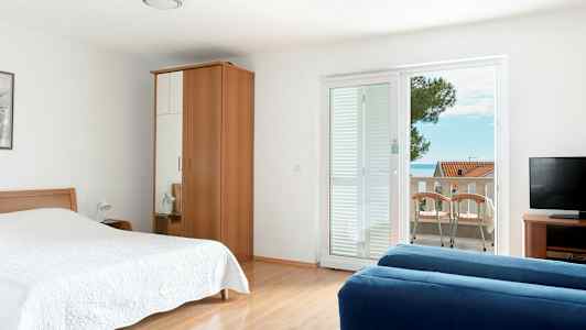 Et hotelværelse med seng, garderobe, blå sofa, fjernsyn og altan med udsigt. (AI-genereret tekst)