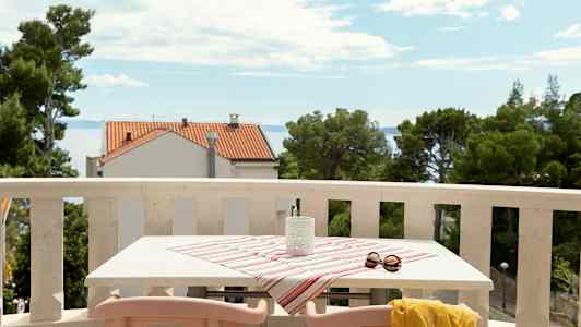 En altan med bord dækket af stribet dug, solbriller og gul sjal, med udsigt over havet og hus. (AI-genereret tekst)