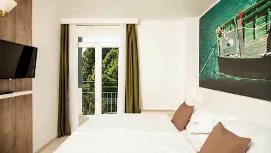 Et hotelværelse med en dobbeltseng, et vægmonteret TV og et stort vindue med grønne gardiner. (AI-genereret tekst)