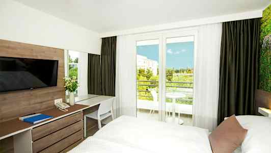 Hotelværelse med to senge, skrivebord og udsigt til altanen og grønne områder. (AI-genereret tekst)