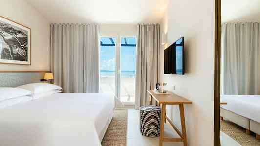 Et hotelværelse med en stor seng, et skrivebord og et TV på væggen. Store vinduer med udsigt til havet. (AI-genereret tekst)
