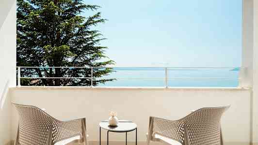 Udsigt fra en balkon med to stole og et bord, mod hav og træ. (AI-genereret tekst)