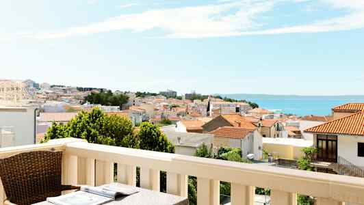 Altan med bord og stol, udsigt over en by med røde tage og havet i baggrunden. (AI-genereret tekst)