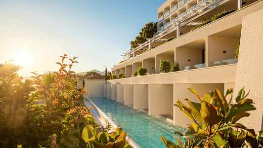 En hotelbygning med en langsgående pool foran, omgivet af planter og solopgang. (AI-genereret tekst)