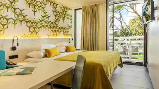 Fotografi af et hotelværelse med et gult sengetæppe og dekorativ vægpanel samt udsigt til et træområde og altan. (AI-genereret tekst)