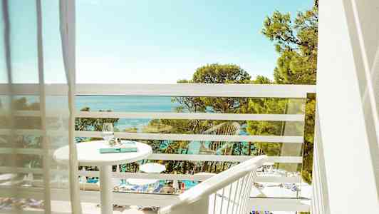 Udsigt fra en balkon over en pool og havet med træer i forgrunden. (AI-genereret tekst)