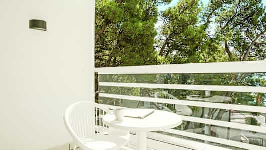 En balkon med et hvidt rundt bord og en hvid stol. Udsigt over grønne træer. (AI-genereret tekst)