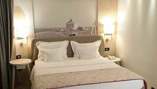 Hotelværelse med dobbeltseng, hvide puder og en vægdekoration med fotografi af et hus på landet. (AI-genereret tekst)