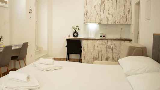 Billede af et lyst indrettet værelse med seng, køkkenbord og bord med stole. (AI-genereret tekst)