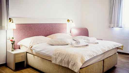 Et værelse med en dobbeltseng redt med tæpper og puder, to sammenrullede håndklæder ovenpå. Et natbord med en vase og en blomst til venstre og et vindue med gardiner til højre. (AI-genereret tekst)