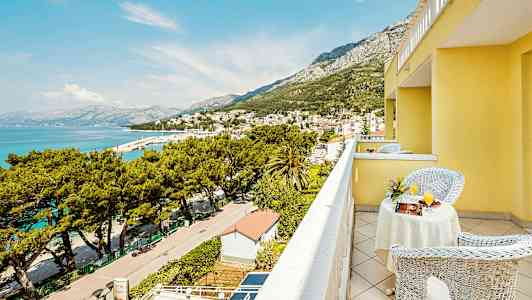 Udsigt fra en balkon på en gul bygning over en by ved havet og omkringliggende bjerge. (AI-genereret tekst)