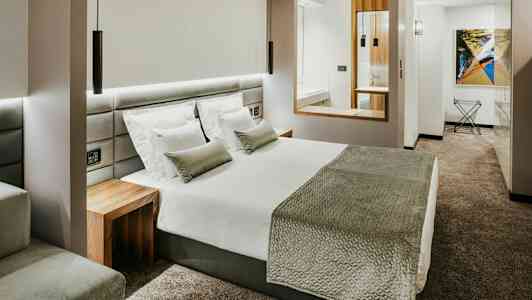 Et moderne hotelværelse med en stor seng, hvide sengetøj, gråt sengetæppe og puder samt gråt væg-til-væg tæppe. (AI-genereret tekst)