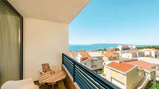 Udsigt fra en altan med to stole, et bord og udsigt over byområdet og havet. (AI-genereret tekst)